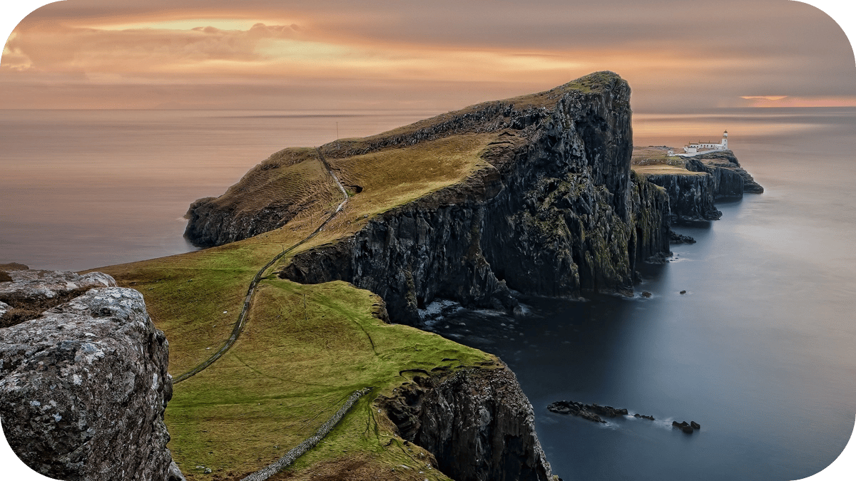 A coastal path on a cliff edge by the sea, with a lighthouse in the far distance.