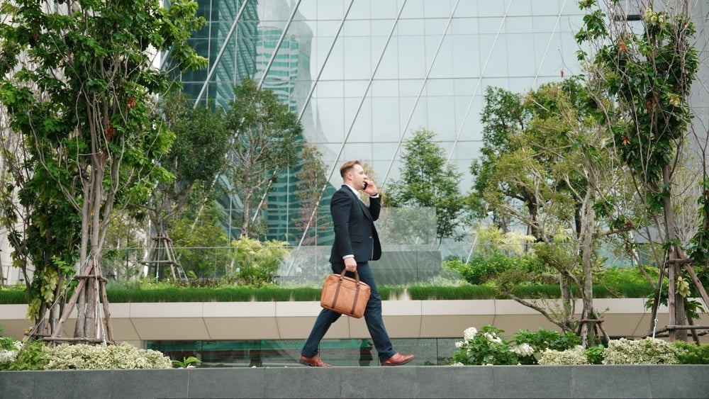 Side view of businessman on phone walking at modern building in green eco city