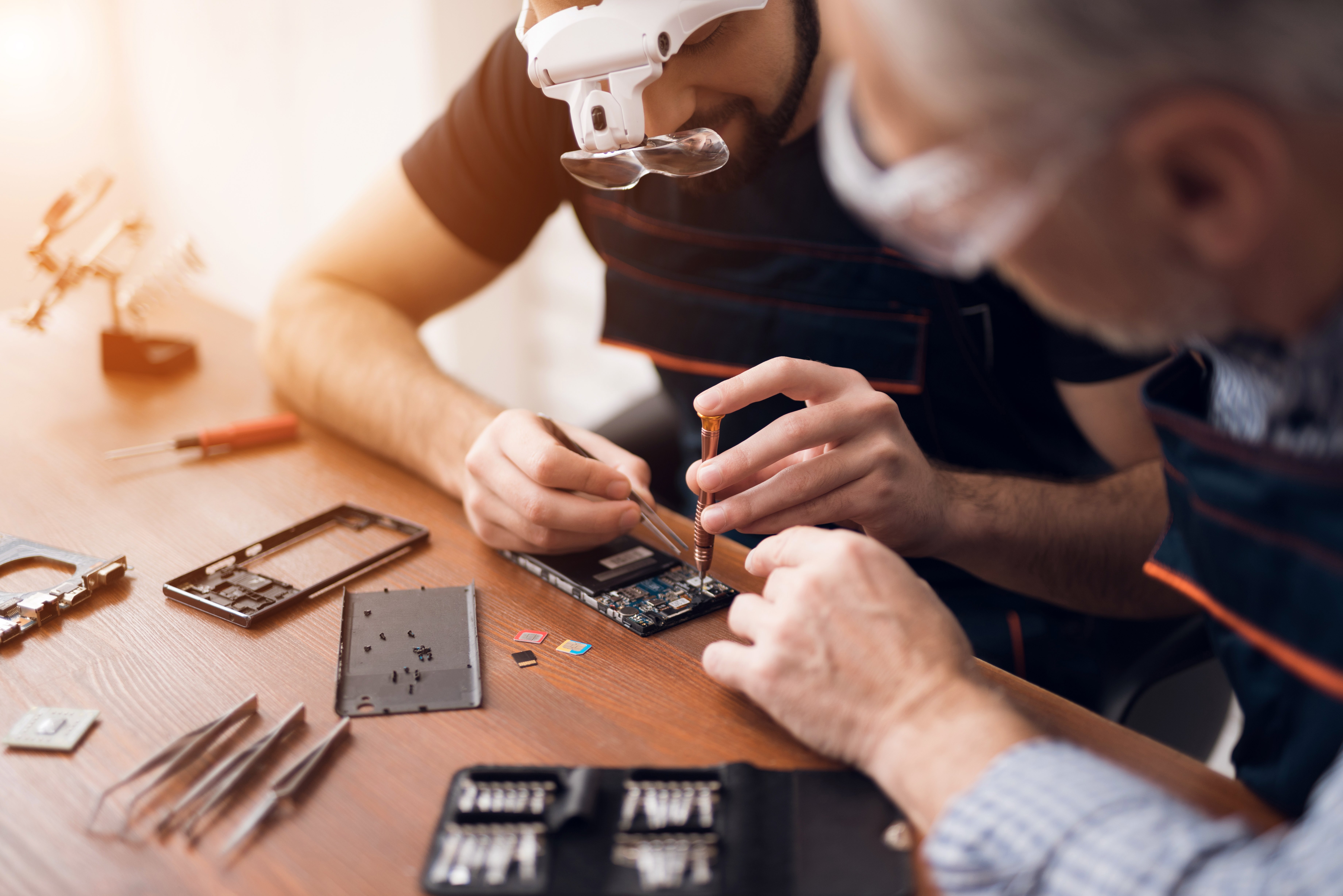 An elderly and young men repair a mobile phone together in a repair shop. 